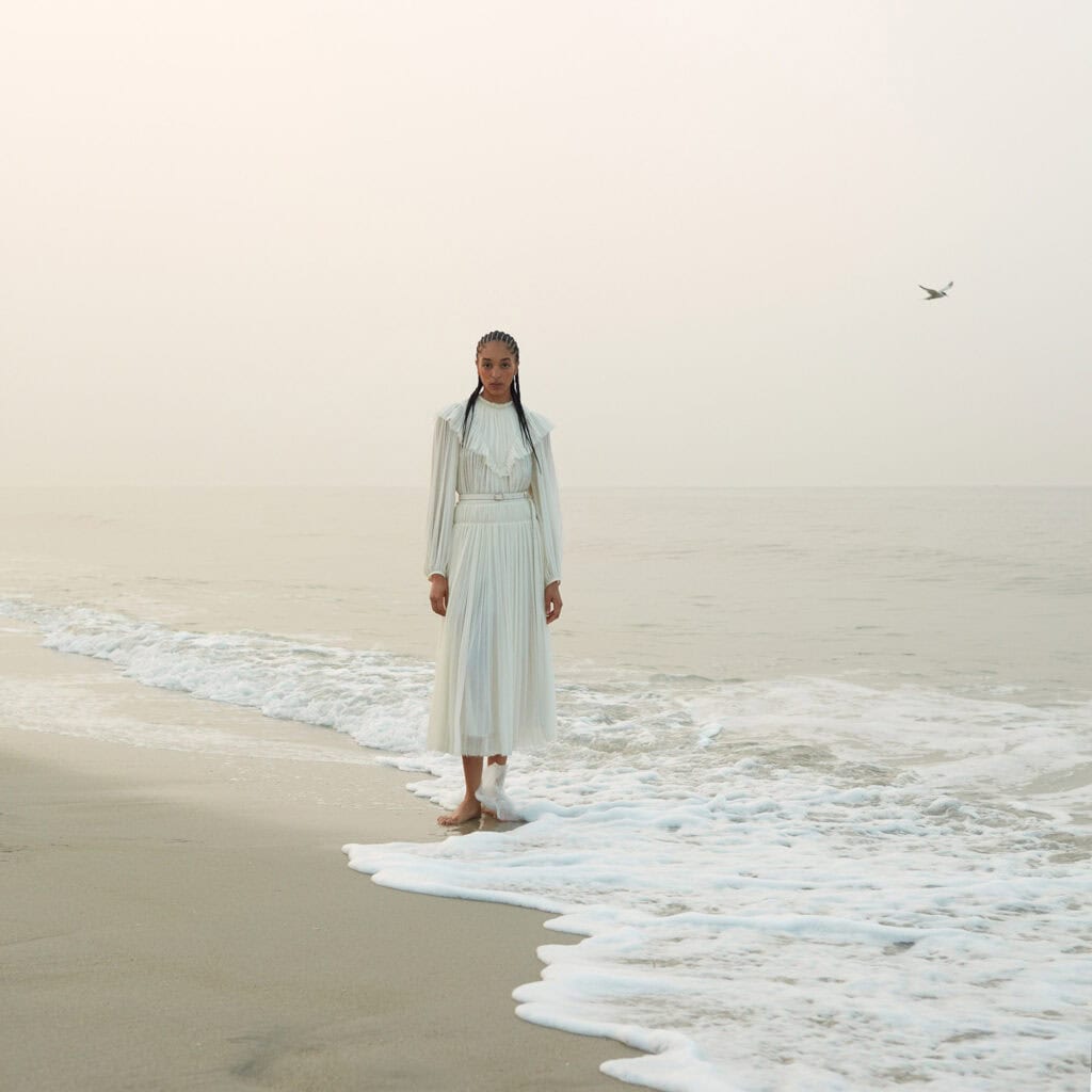 Woman in white dress standing on the beach with ocean waves and a bird in the sky.