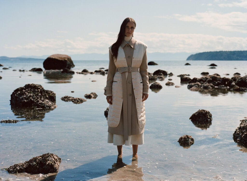 Elegant woman standing in shallow ocean water with rocks, showcasing modern feminine style.