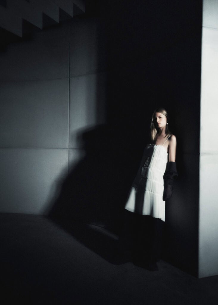 Model in a white dress standing against a dark wall with shadow effects.