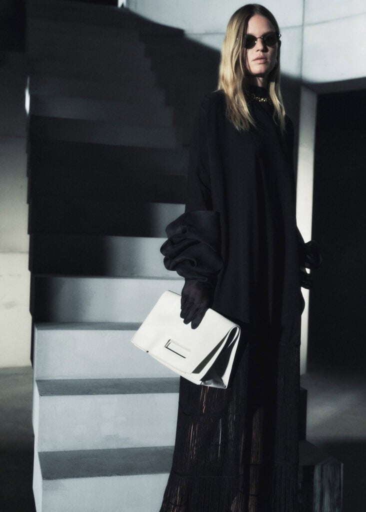 Model wearing KHAITE Anna 2023 collection, holding a white handbag, in a modern indoor setting.