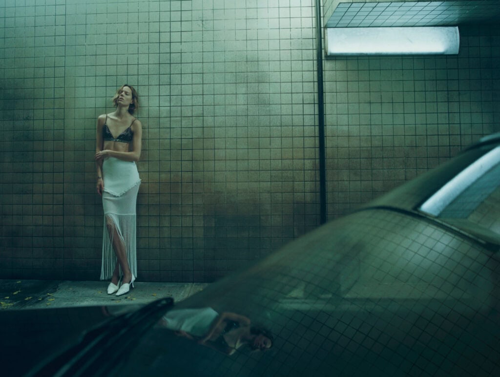 Model in KHAITE Freja dress standing against tiled wall in urban setting.