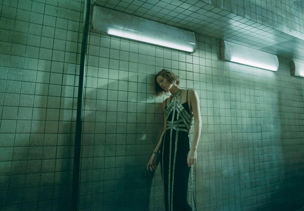 Model wearing KHAITE Freja 2022 in an urban setting with tiled walls.