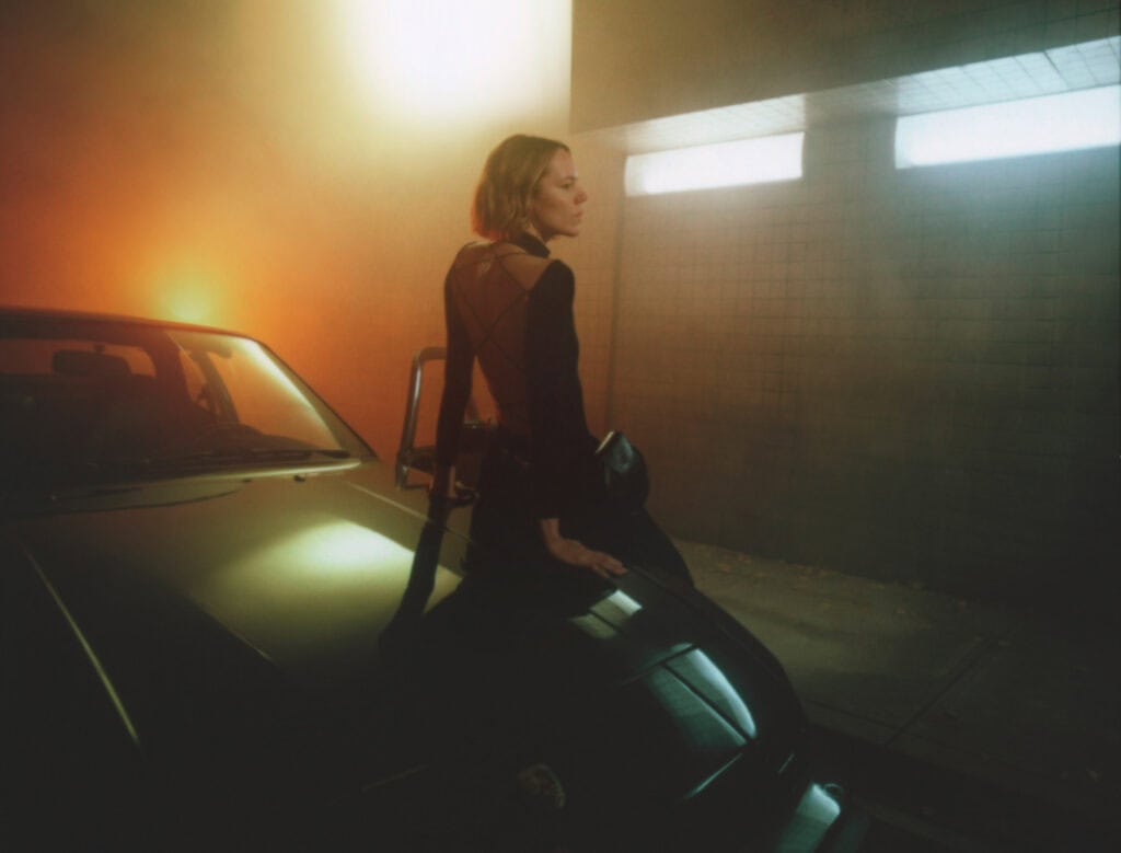 Fashion model standing beside a vintage car in a dimly lit garage.