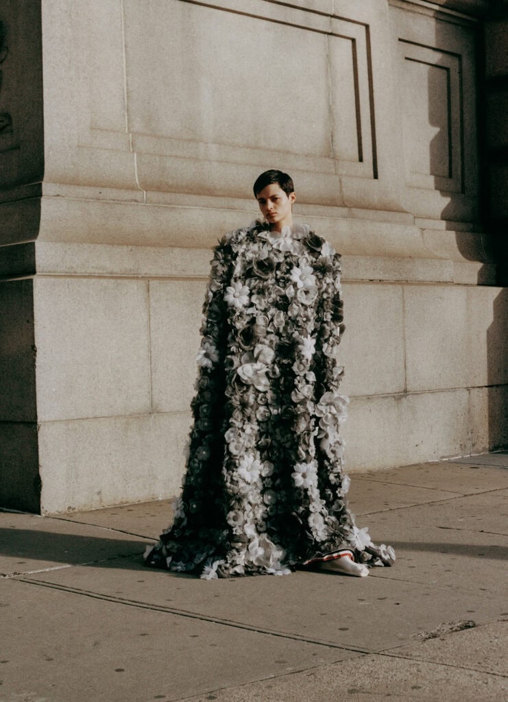 Fashion model in a floral dress standing against a stone building exterior.