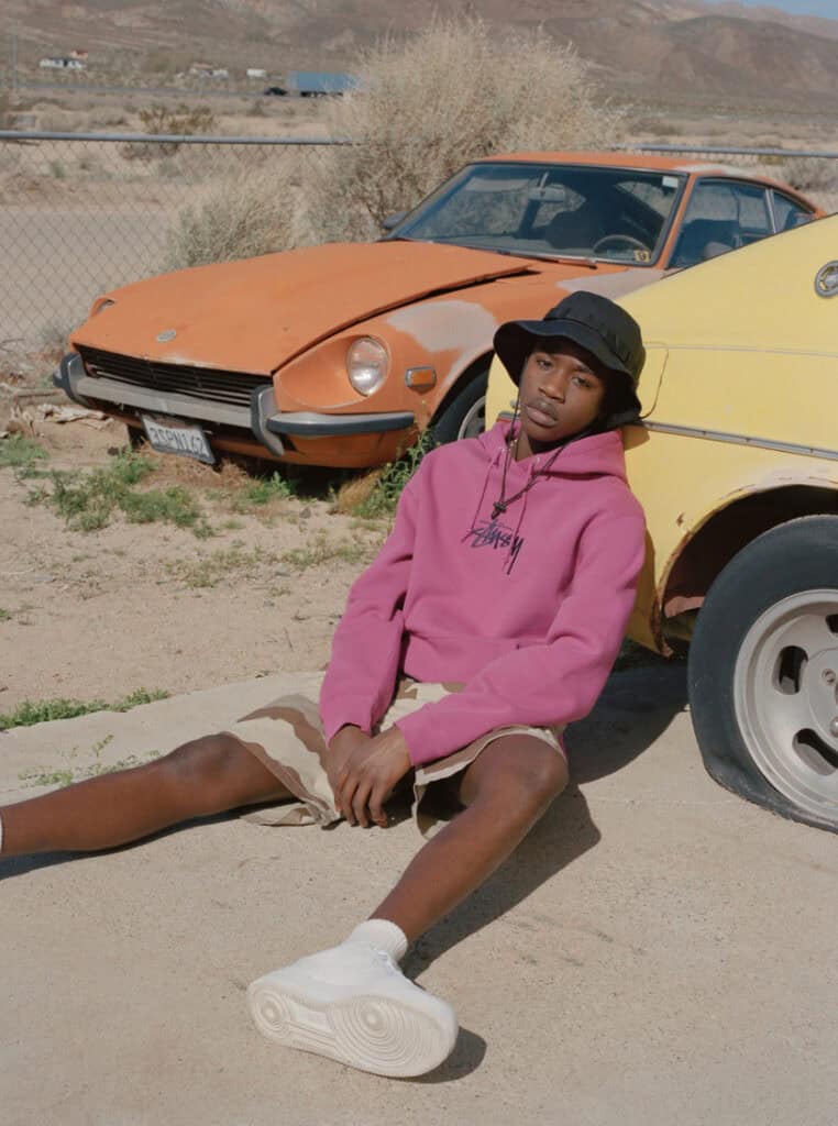 Vintage cars in desert landscape with a young man in pink hoodie sitting on the ground.