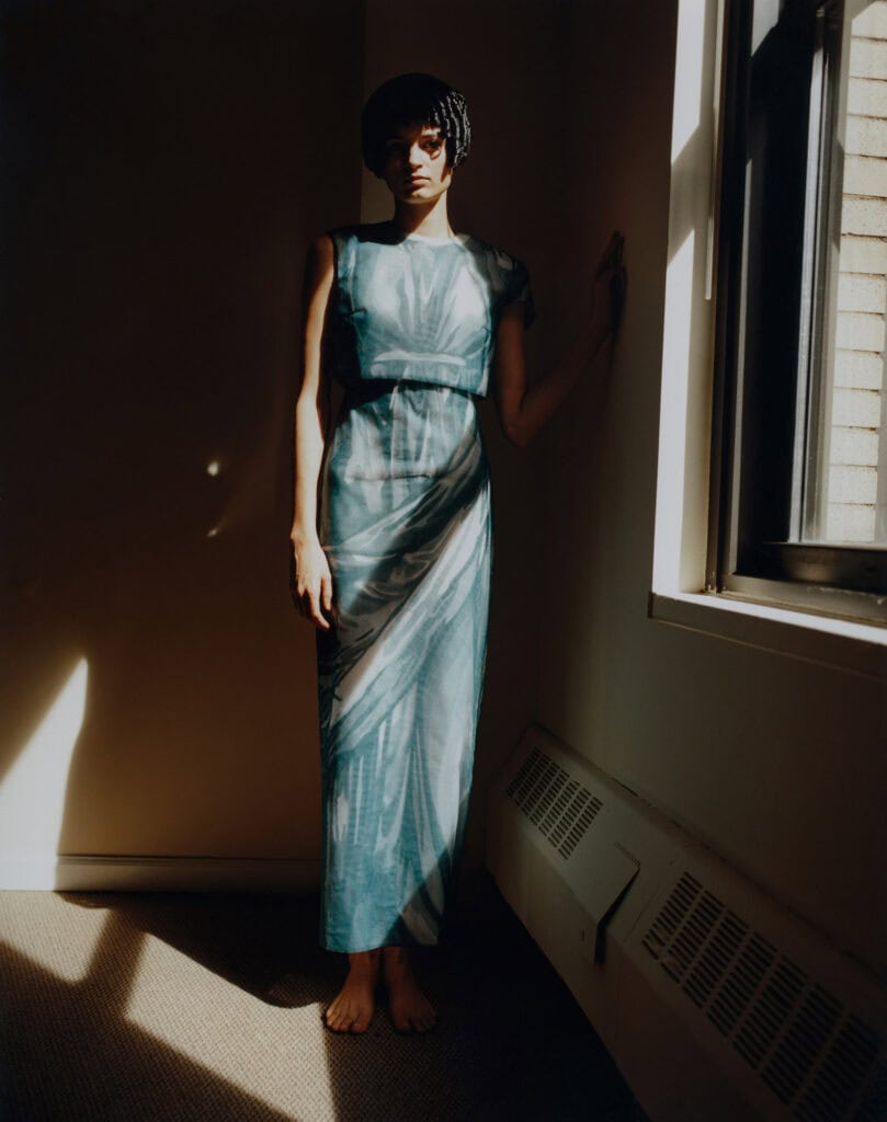 Elegant model in a flowing blue dress by Thom Brown, standing by a window with natural light.