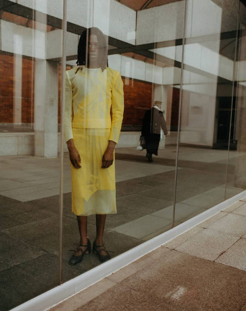 Image showing a model in a yellow dress reflected in a glass window at a fashion event.