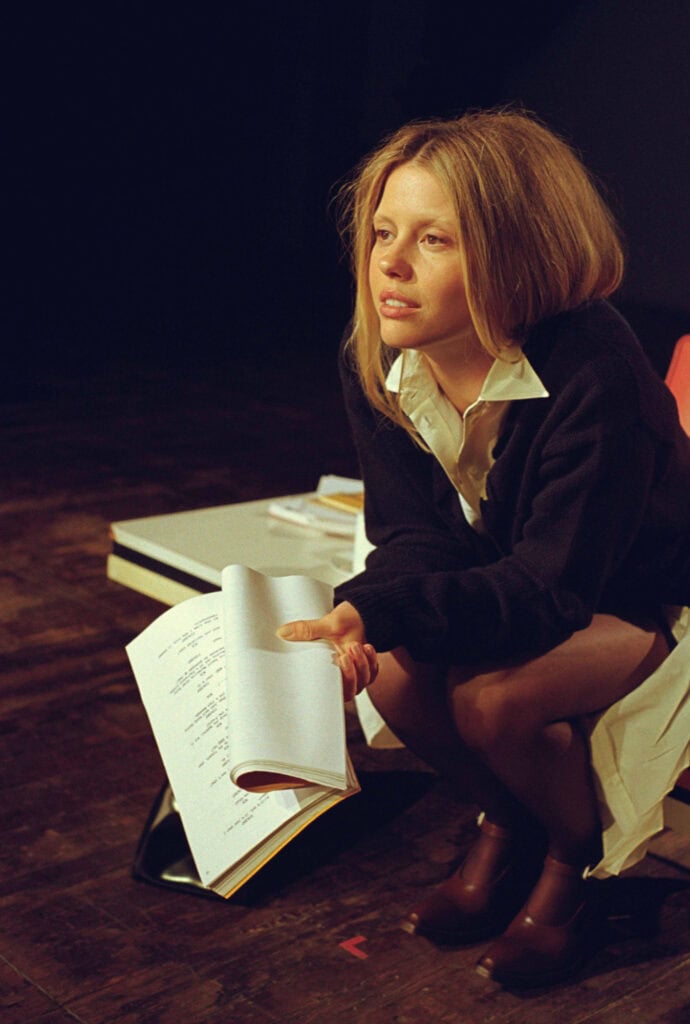 Young woman with shoulder-length hair reading a script on stage during a performance.