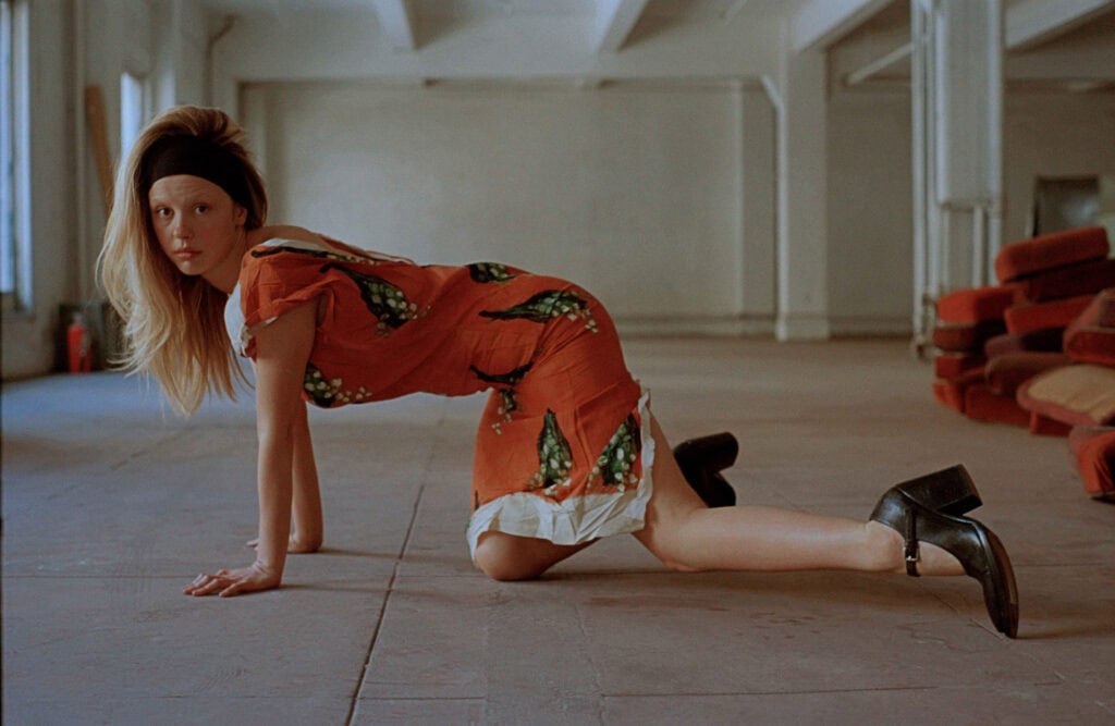 Mia Goth in a striking pose on the floor, wearing a vibrant orange dress with green patterns, in an.