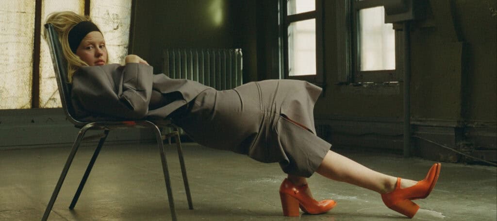 Mia Goth reclining on a chair in a dimly lit room, wearing a gray outfit and orange heels.