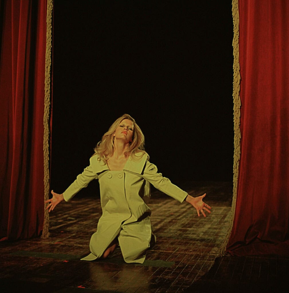 Mia Goth performing on stage with open arms, dressed in a light-colored outfit, in front of red curt.